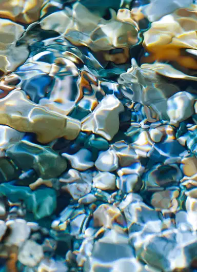 A close-up view of colorful, smooth rocks submerged in clear water, with sunlight creating shimmering reflections and ripples on the surface.